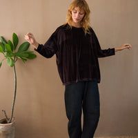 Woman wearing a dark brown velvet top and black pants standing next to a plant against a beige wall.