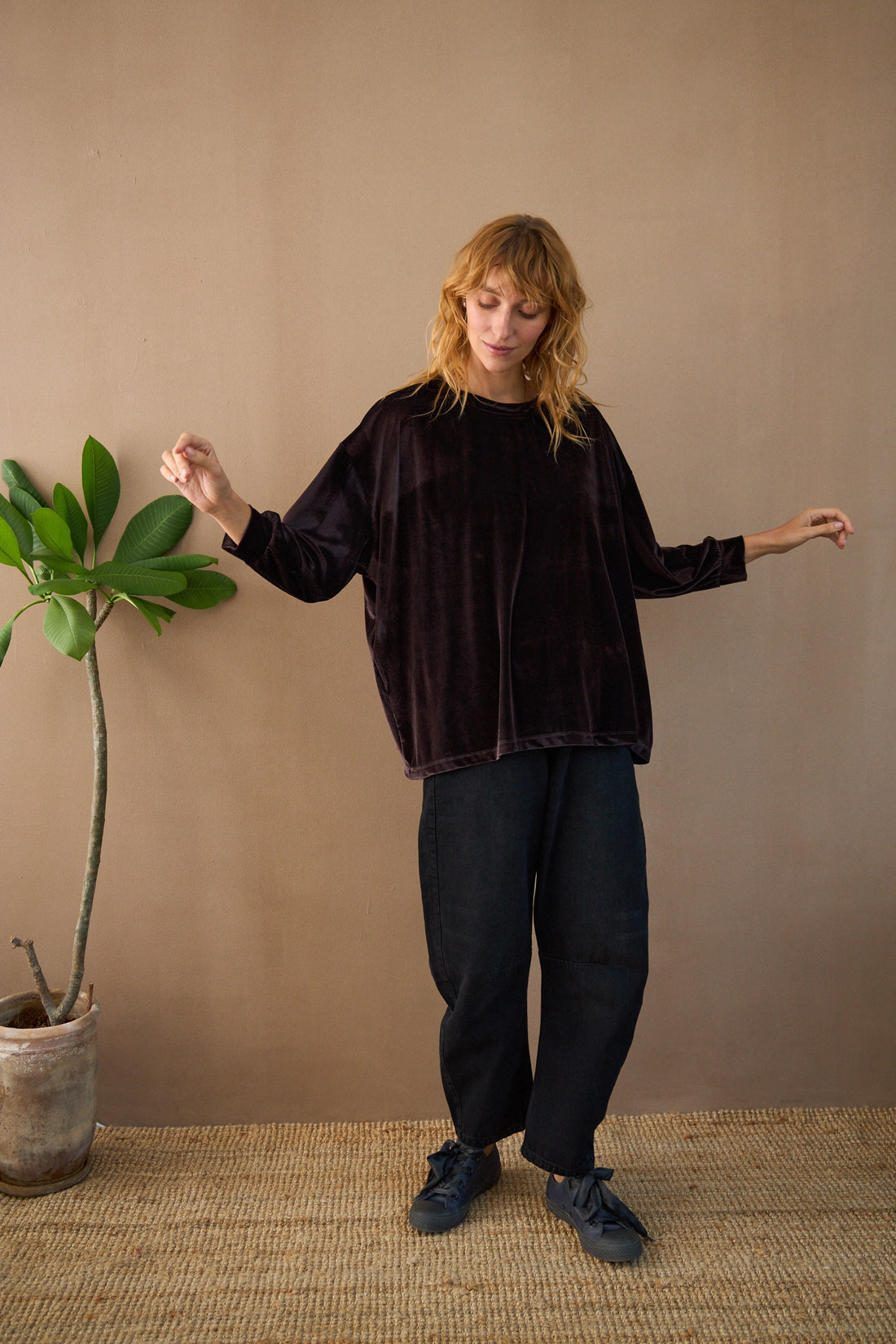 Woman wearing a dark brown velvet top and black pants standing next to a plant against a beige wall.
