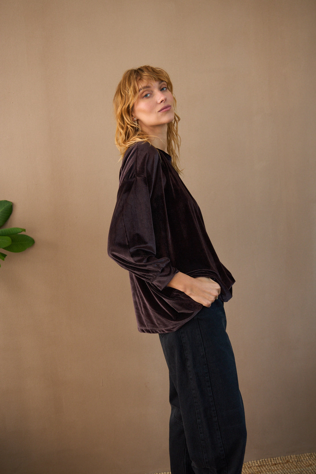 Woman wearing a dark brown long-sleeve top and black pants against a beige wall.