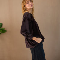 Woman wearing a dark brown long-sleeve top and black pants against a beige wall.
